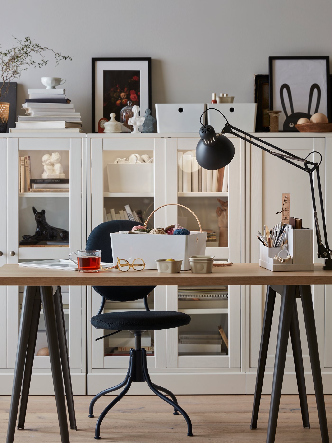 Tablero con una lámpara de trabajo, un organizador de escritorio, una taza y una silla negra. Al fondo hay una combinación de almacenaje con puertas de vidrio.