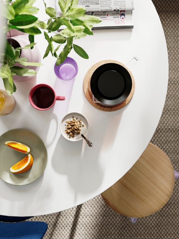 Une table DOCKSTA dressée pour le déjeuner avec du café, des fruits et des bols, aux côtés d’un tabouret avec une plante en pot en arrière-plan.