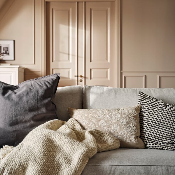 A MURSENAP cushion and cushions with a beige/black KUSTFLY cushion cover and a dark grey DYTÅG cushion cover lie on a sofa.