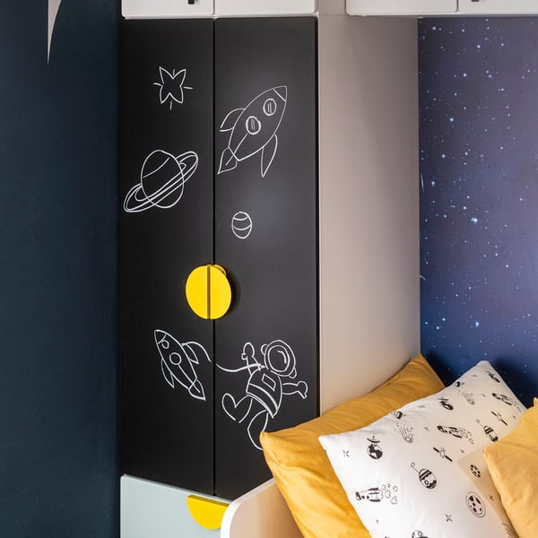 Un armadio SMÅSTAD con ante nere a lavagna e maniglie BEGRIPA gialle con accanto dei cuscini colorati.