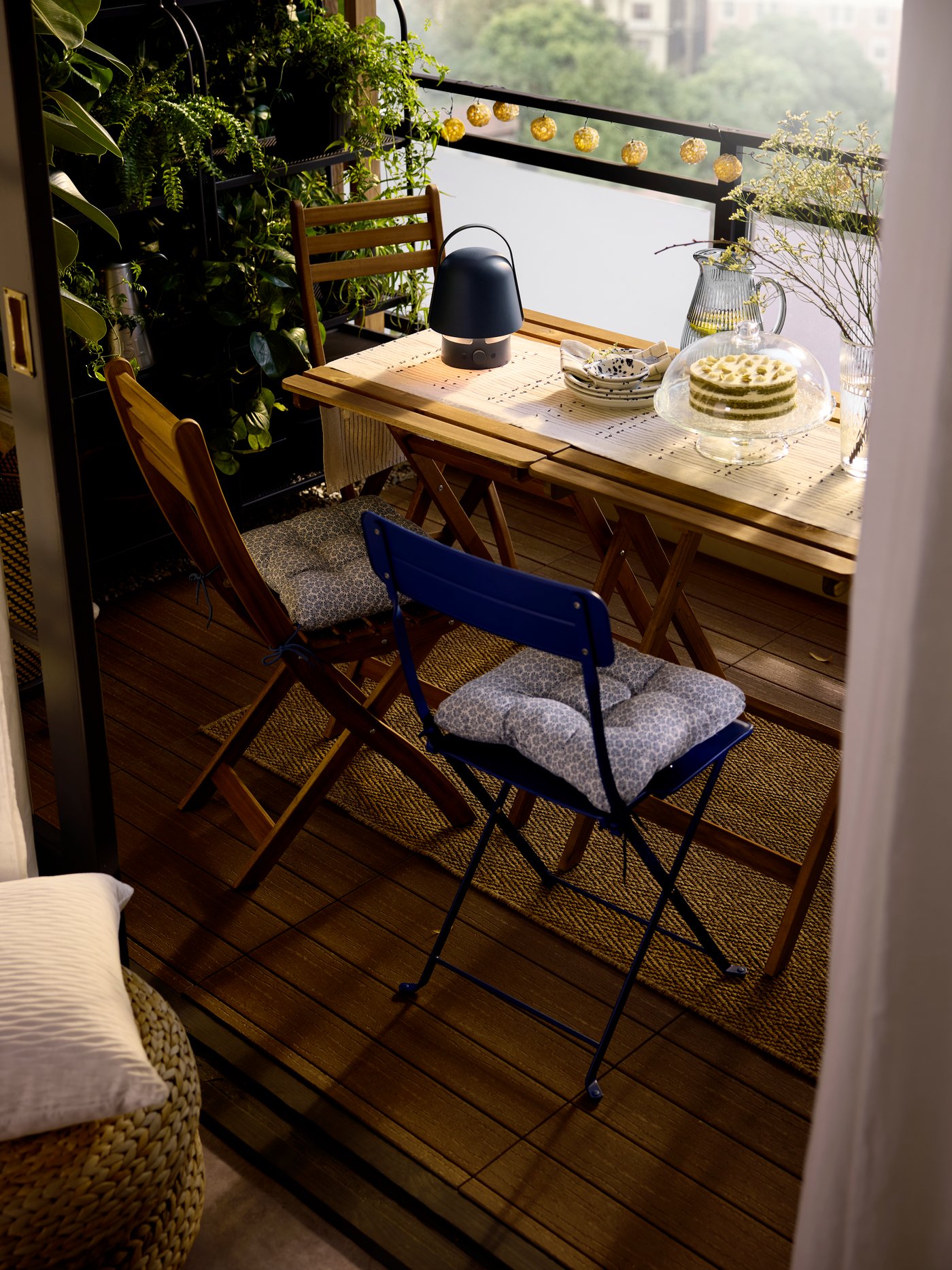 A lush balcony with small chairs and tables, holding a blue outdoor VAPPEBY Bluetooth speaker lamp, plates and a cake stand.