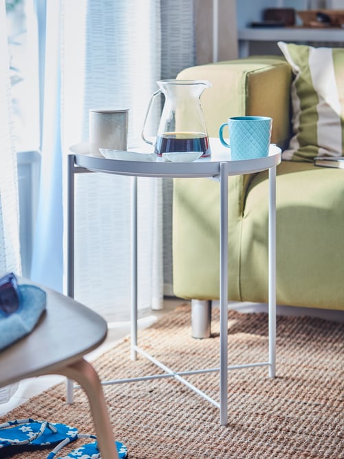 Living room with white tray table, two-seat sofa, cushion cover, blue mug, coffee maker, a pair of curtains, flatwoven rug.