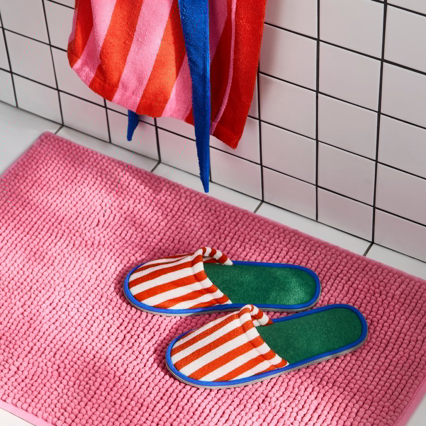 Two SKOGSSALLAT slippers with red and white stripes on the outside, green and blue on the inside. Pink bath mat, white floor.