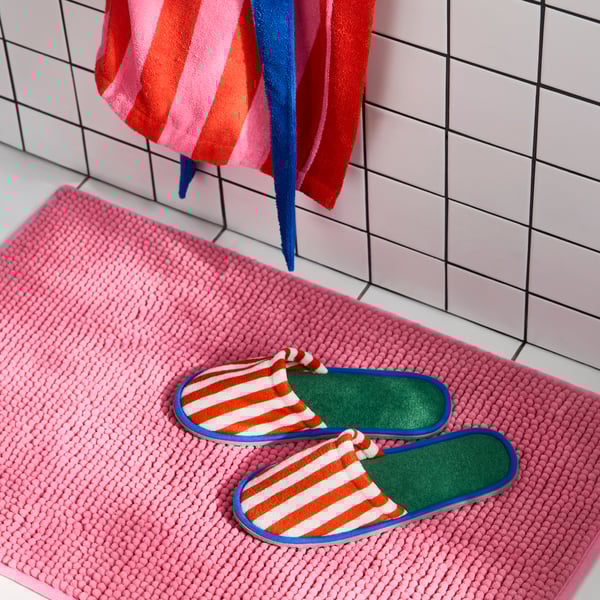 Dues sabatilles SKOGSSALLAT de ratlles vermelles i blanques a fora, i de color verd i blau a dins. Estoreta de bany rosa en un terra blanc.