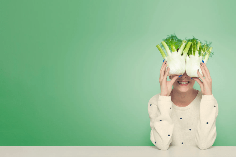 Une personne assise à une table blanche devant un fond vert, portant des branches de céleri frais sur la tête, dans une mise en scène originale.