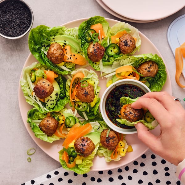 A hand has taken a meatball wrapped in a lettuce leaf from a pink plate and is dipping it into a white dish with soy sauce.