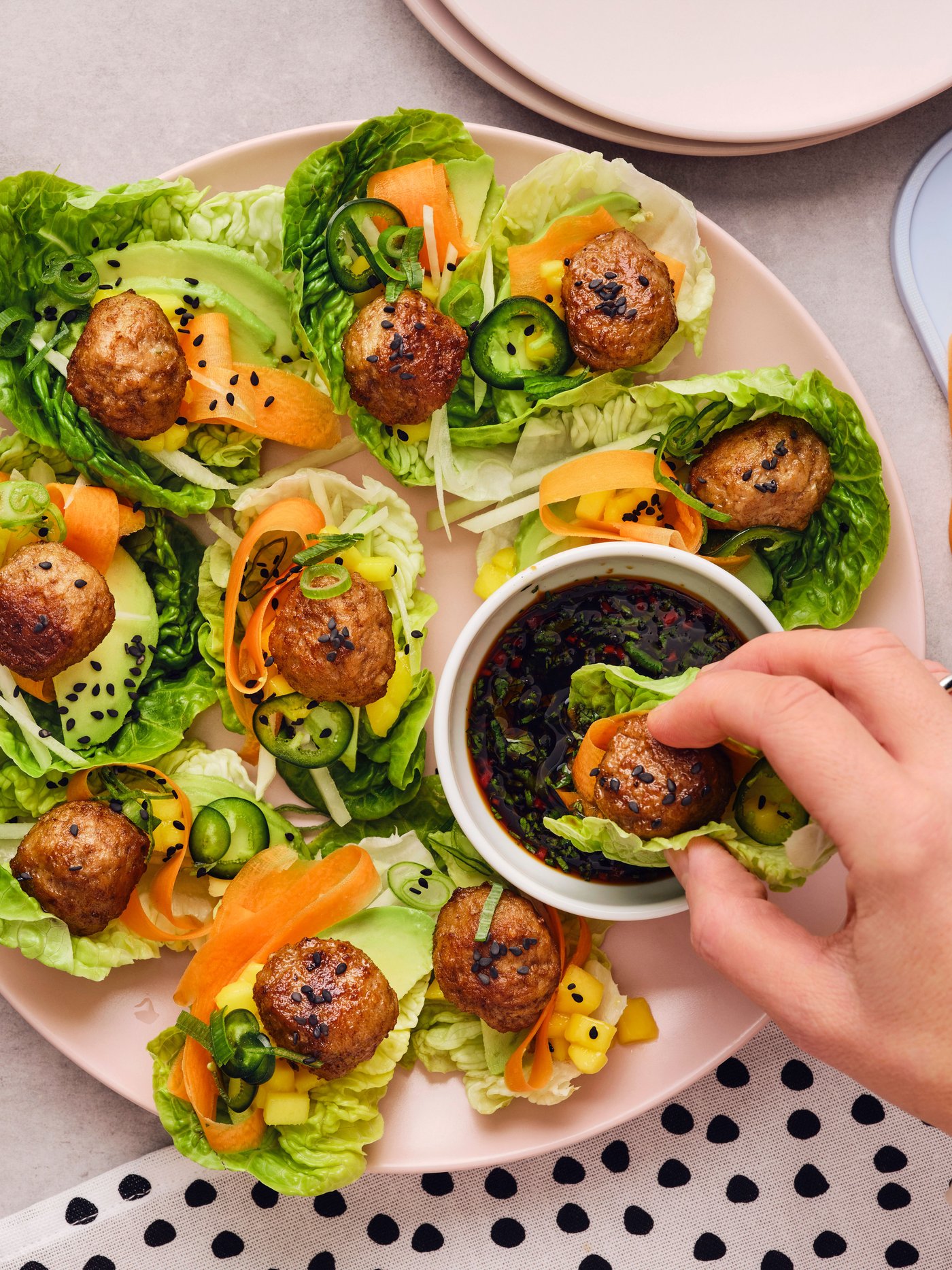 A hand has taken a meatball wrapped in a lettuce leaf from a pink plate and is dipping it into a white dish with soy sauce.