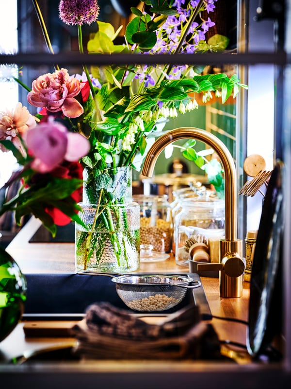 A kitchen sink with a brass DELSJÖN kitchen mixer tap with a vase of fresh pink and purple flowers on the counter.