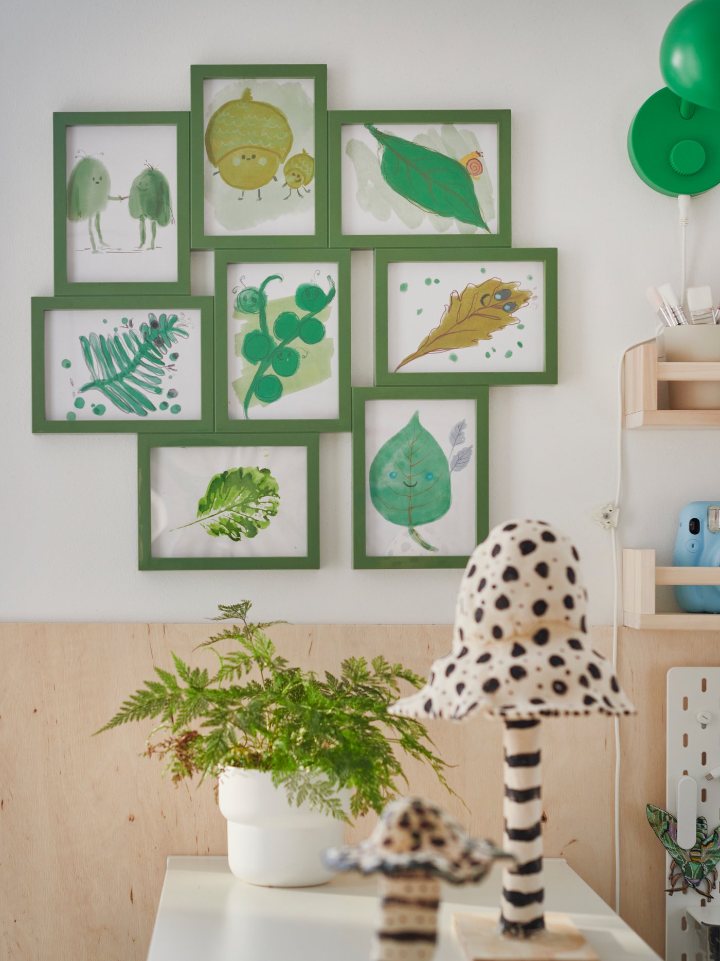 Une pièce où un cadre collage VÄXBO vert accueille huit dessins disposés sur un mur blanc, au-dessus d'étagères remplies de plantes.
