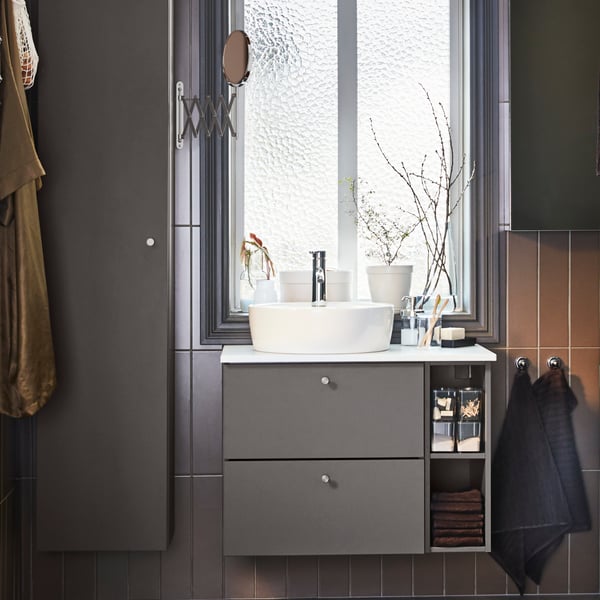 Un bagno in stile moderno con mobili grigi e lavabo da appoggio. C'è un'ampia finestra da cui inetra luce. A lato, uno specchio in acciaio FRÄCK.