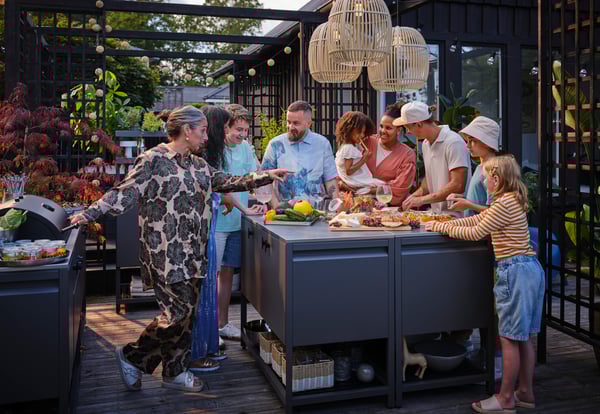 Auf diesem Bild kann man viele Personen sehen die zusammen lachen und an einem hohen schwarzen Tisch stehen. Sie Essen und trinken zusammen und im Hintergrund sieht man bunte Pflanzen und einen Grill.