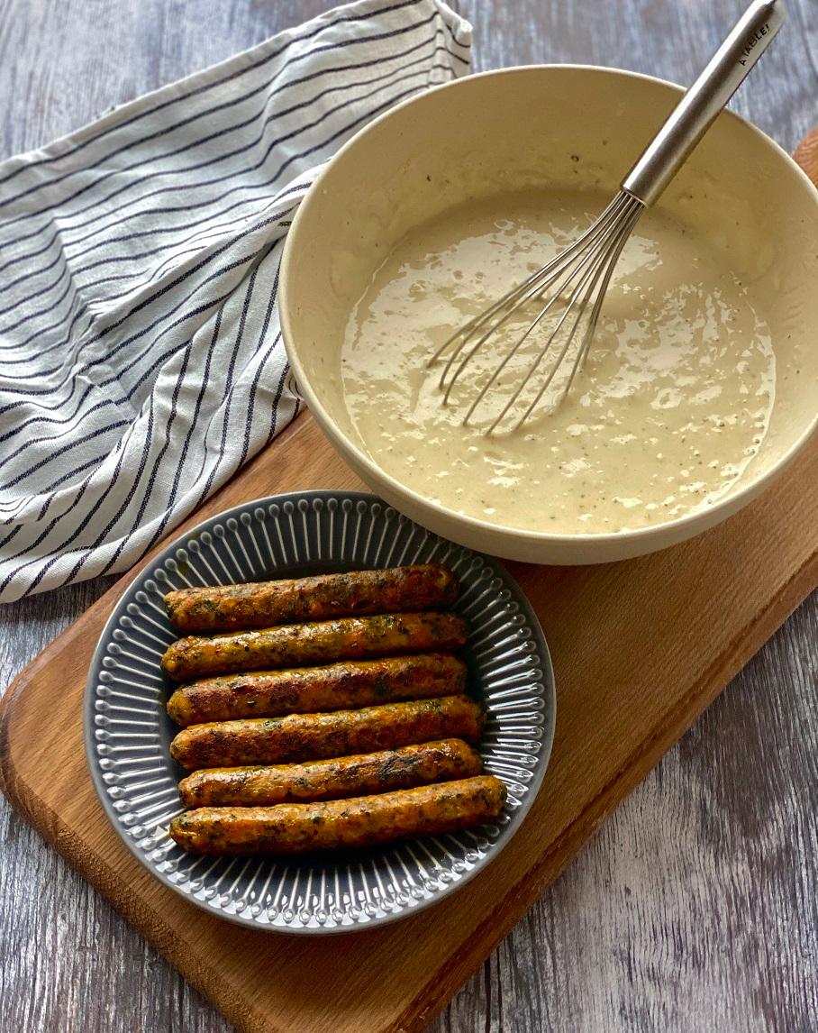 Sur une planche en bois, il y a une assiette avec des saucisses à côté d'un saladier avec un fouet et un torchon rayé en arrière-plan.