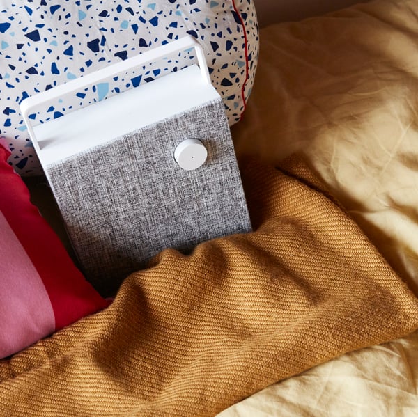 A teen room with a white, portable ENEBY bluetooth speaker standing on a bed with yellow bed linen and cushions.