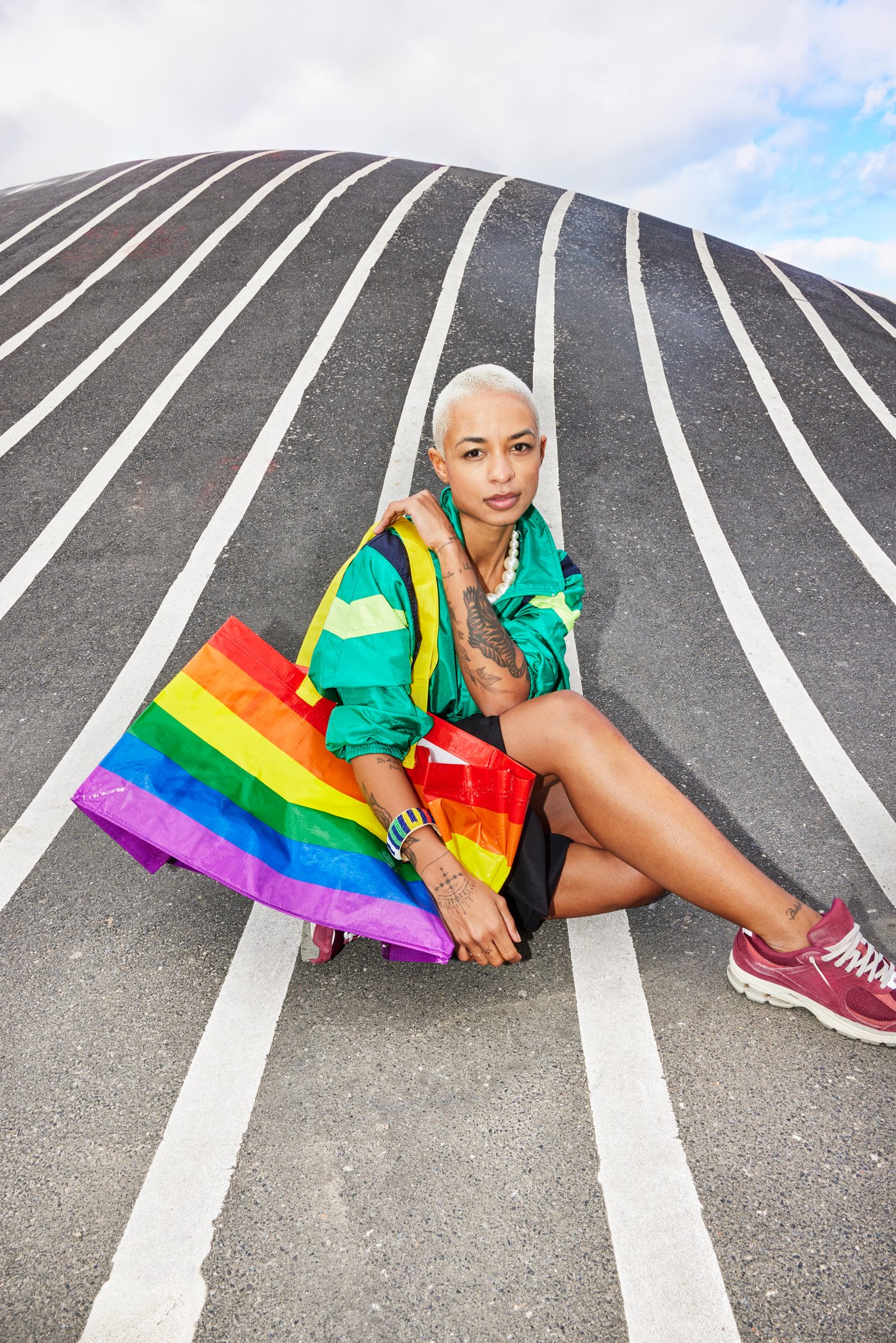 A girl sits on a gray and white striped road with a multicolor STORSTOMMA bag draped on her shoulder.