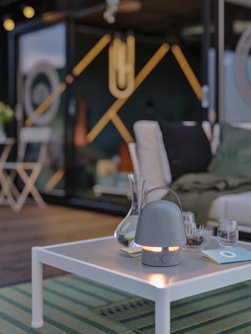 Un spațiu în aer liber care afișează o lampă cu difuzor Bluetooth VAPPEBY lângă o carafă, așezată pe o măsuță de cafea în aer liber.
