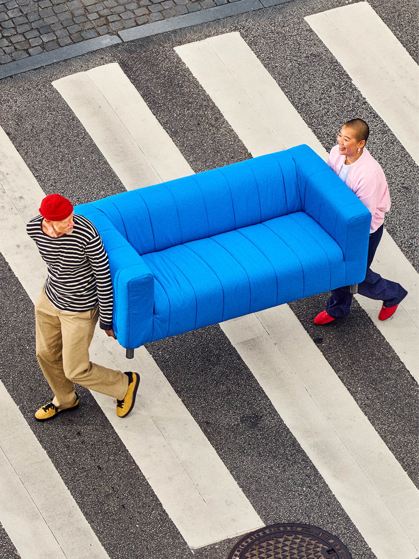 Two people, one in a red beanie and striped shirt and the other in a pink shirt, carefully carry a bright blue IKEA KLIPPAN sofa across a zebra crossing on a city street, with a manhole cover visible in the foreground.