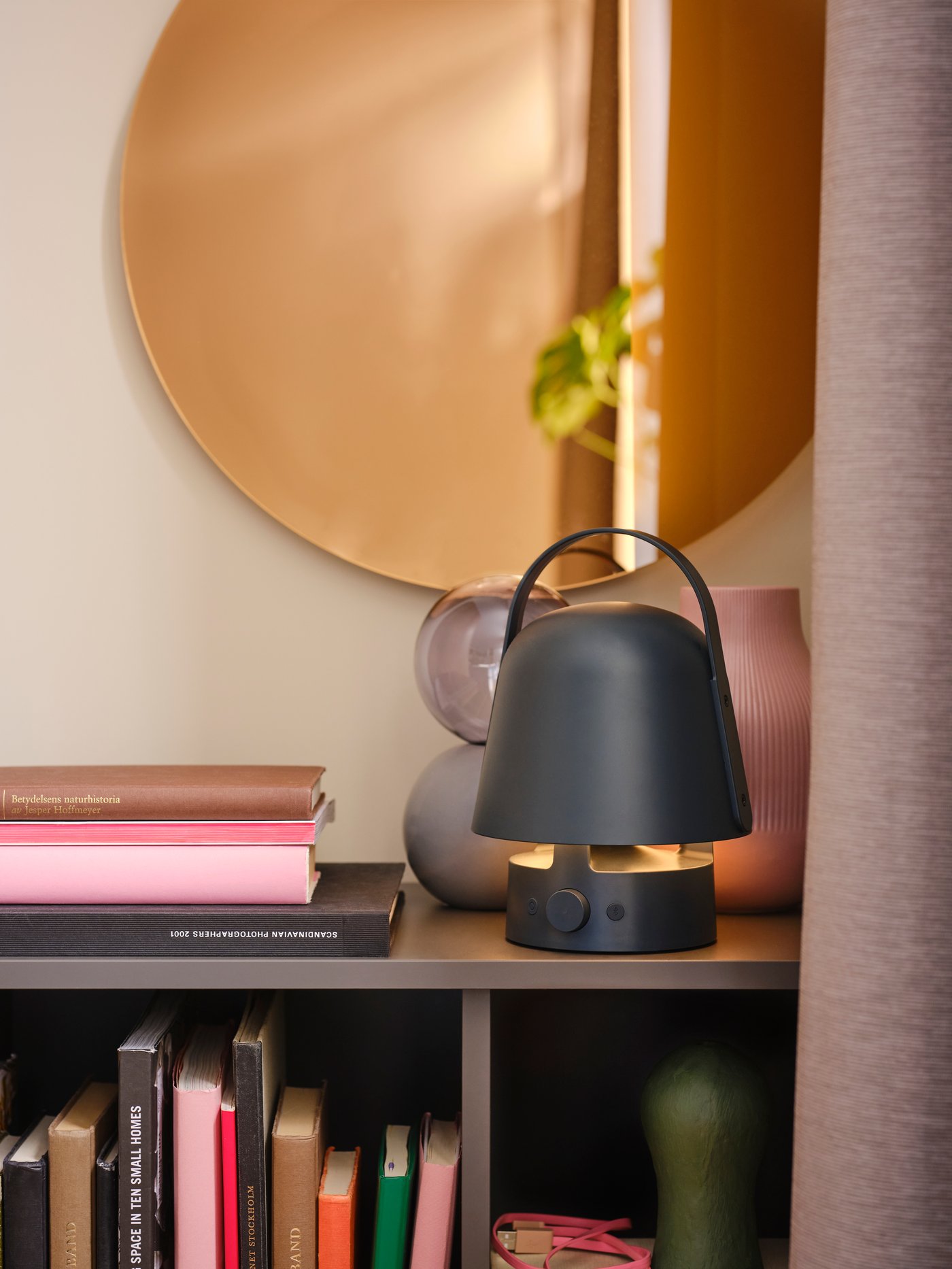 A round, bronze-coloured VÄRMBLIXT LED wall/mirror lamp hangs above a cabinet with decorative items and a speaker lamp on it.