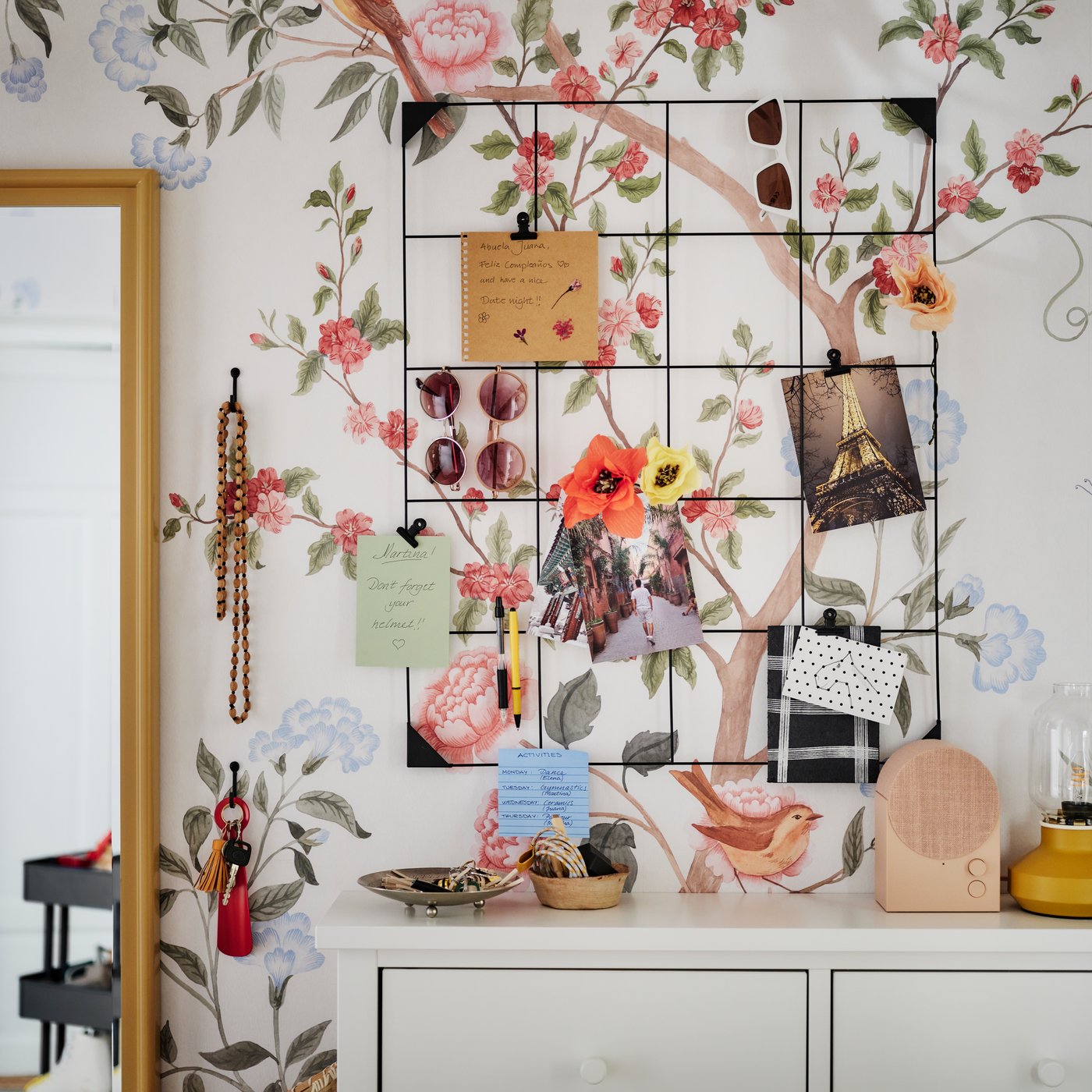 En sort SÖSDALA opslagstavle monteret på en blomstret væg over et hvidt GULLABERG skoskab med kort og solbriller.