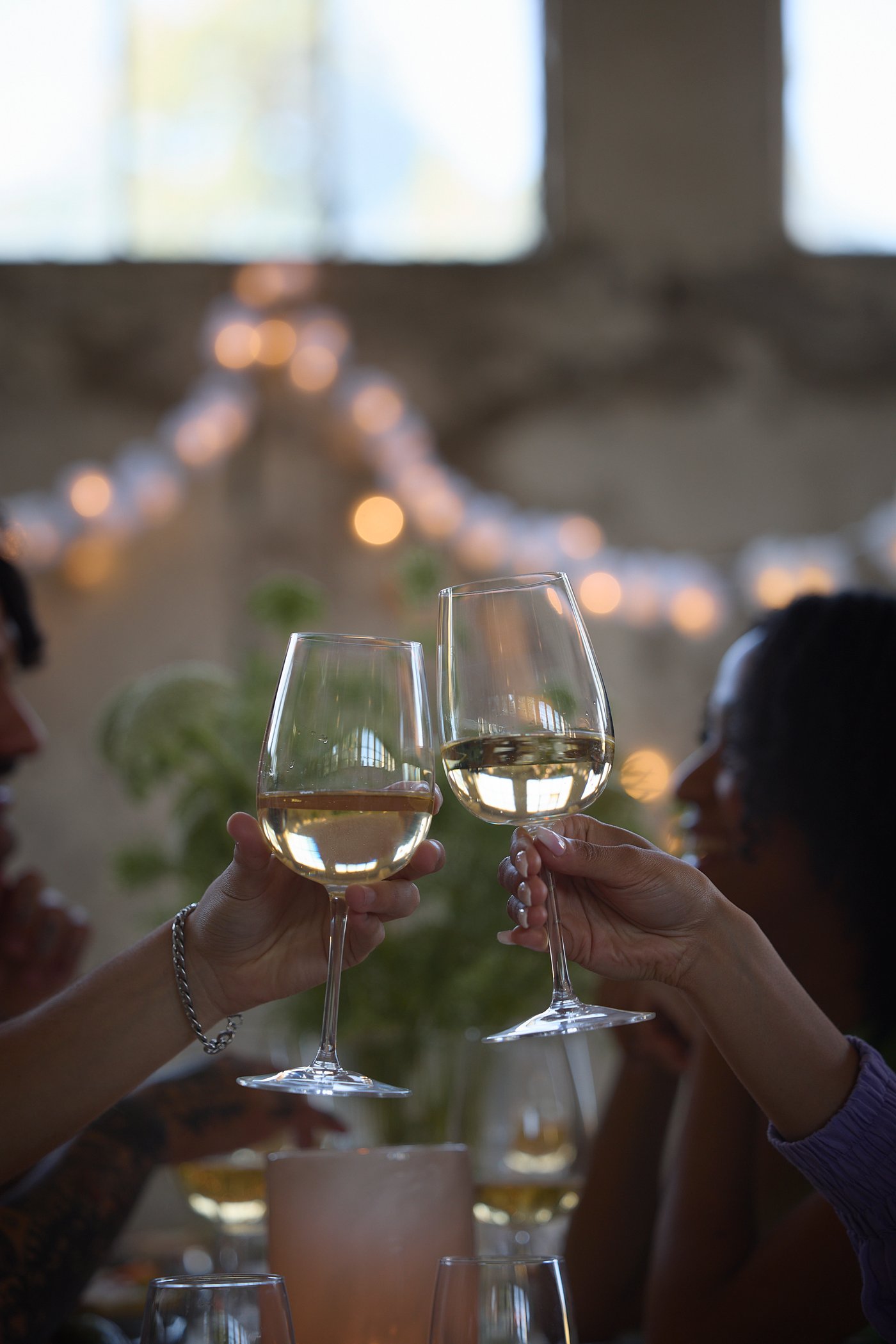 Two people toast with each other in wine glasses from IKEA with string lights in the background.