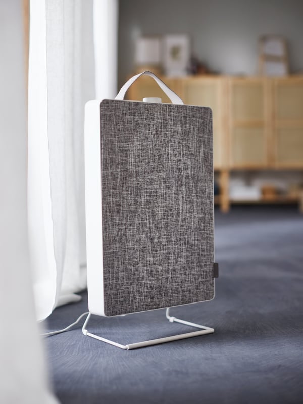 A white FÖRNUFTIG air purifier with a gray front standing next to a white curtain with a row of cabinets in the background.