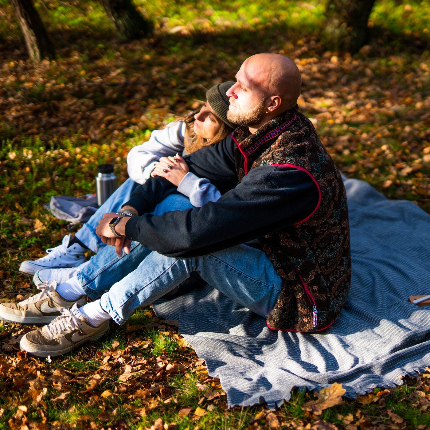 A man and a woman sitting on a picnic blanket in the woods covered by dried autumn leaves.