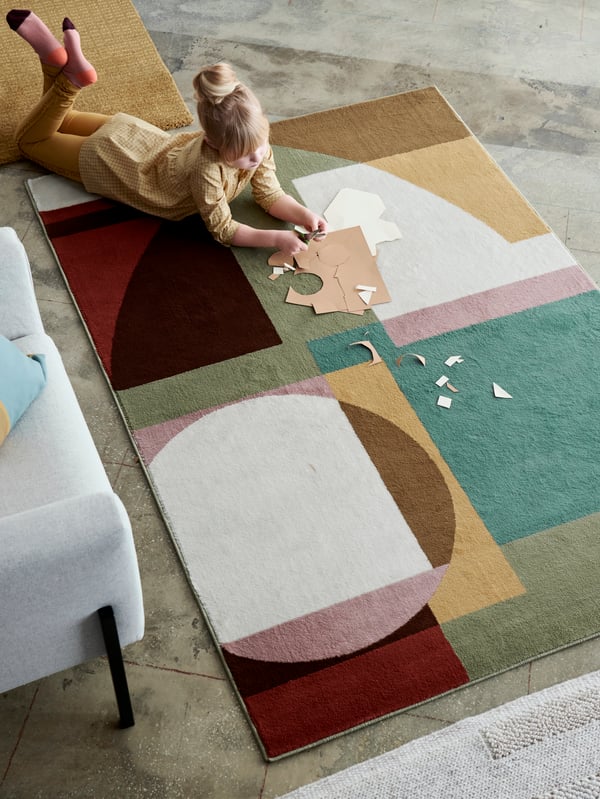 A large, multicolored STENMÄTARE rug with low pile and a bold, geometrical pattern and a girl cutting paper.
