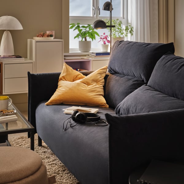 A living room featuring a black-blue MANNARP two-seat sofa with cushions, placed near a table and a window with plants.