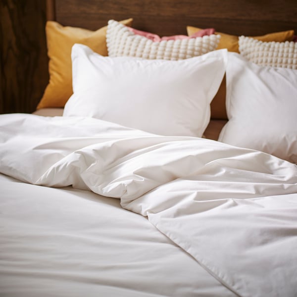 A bed made up with HAMPFLOCKEL bed linen with the duvet folded back and bright colour cushions behind the pillows.