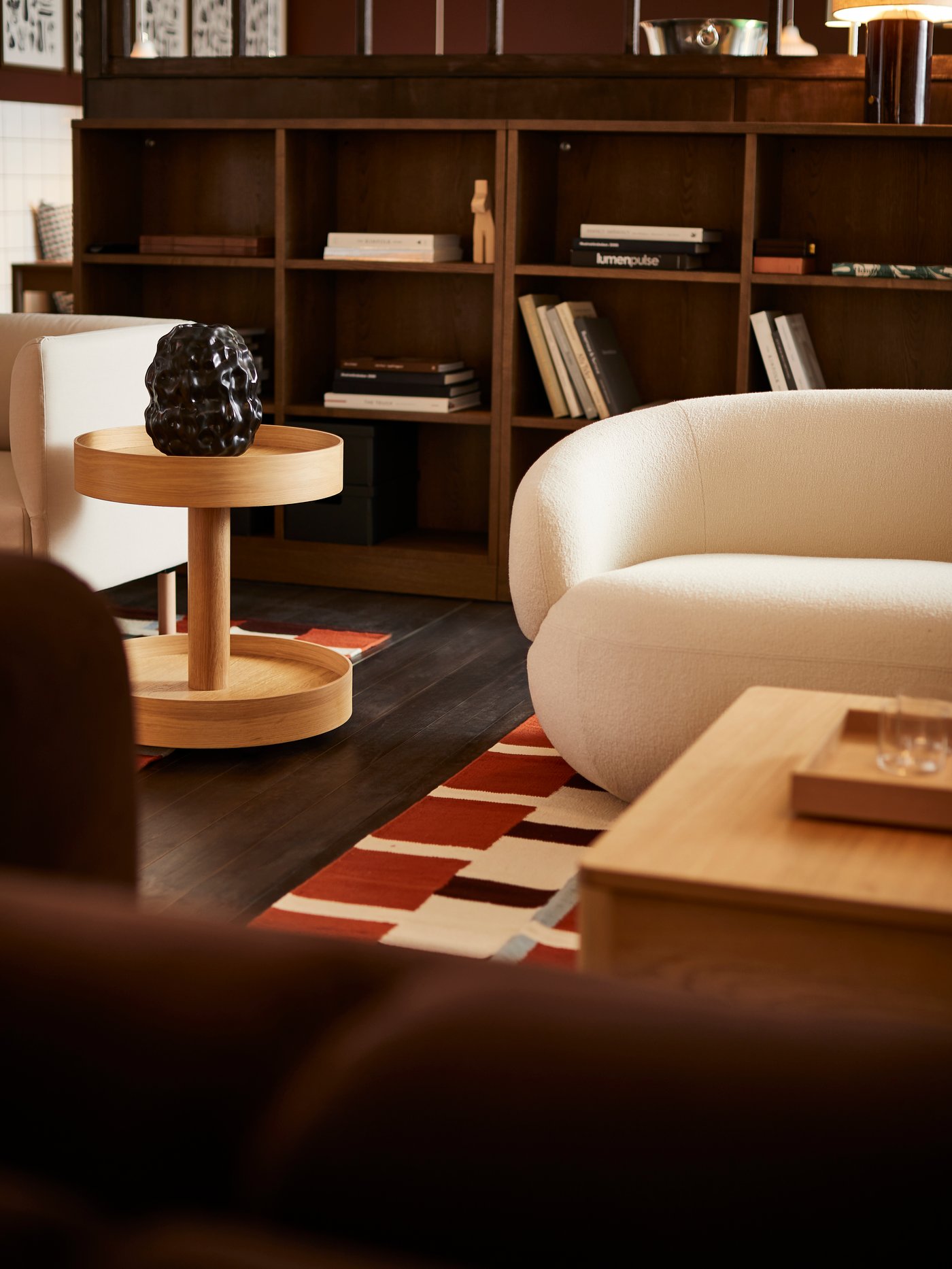 Vardagsrum med vit rundad soffa, träbord och hyllvägg fylld med böcker och dekorationer. Färgstark matta med rött och vitt rutmönster ger kontrast mot mörkt golv.
