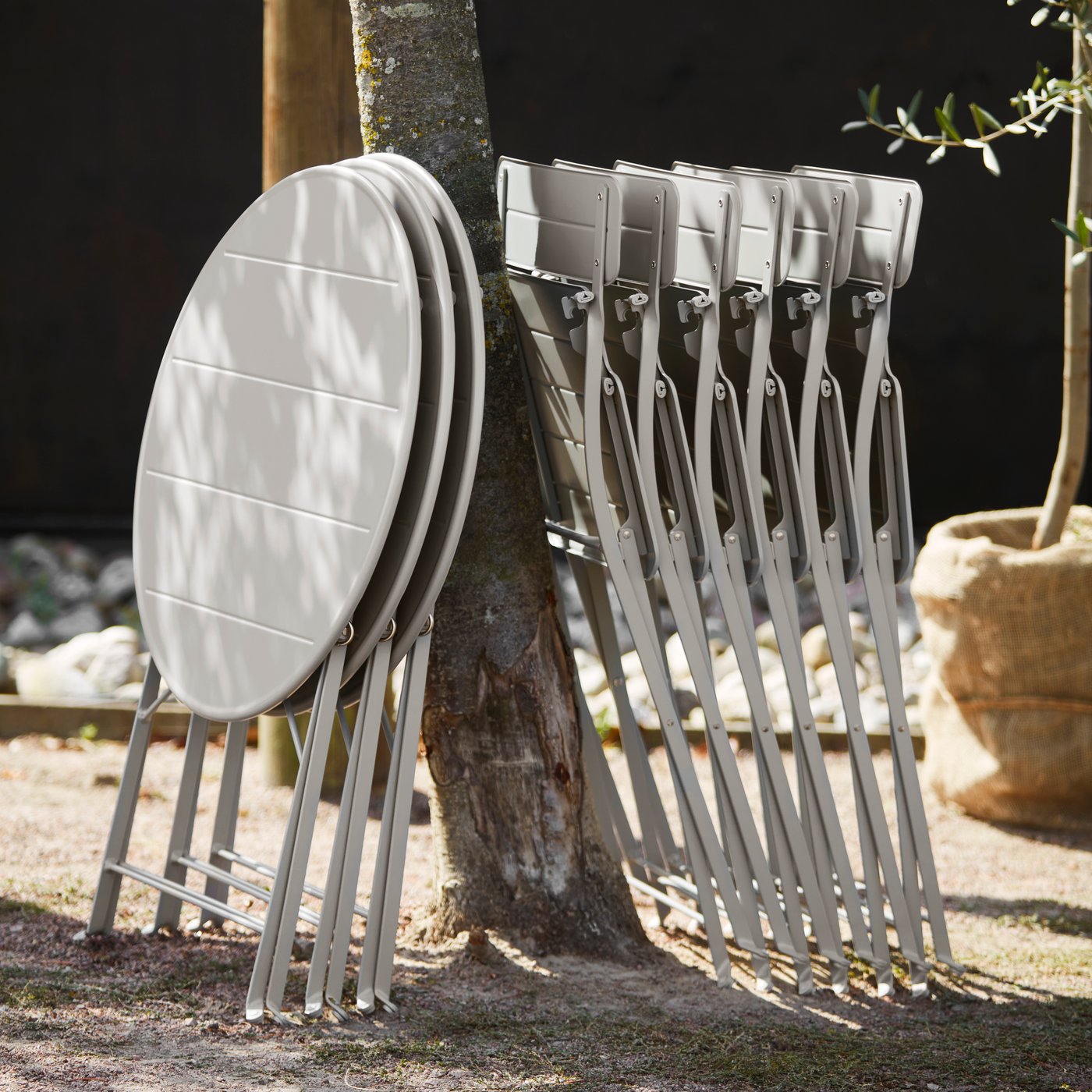 Several folded, off-white SUNDSÖ outdoor chairs and tables are leaning neatly against a tree.