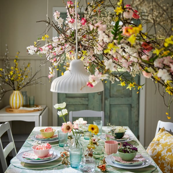 Some colourful SMÖRFISK mugs and UPPLAGA bowls sit on a beautifully set dining table adorned with flowers and greenery.