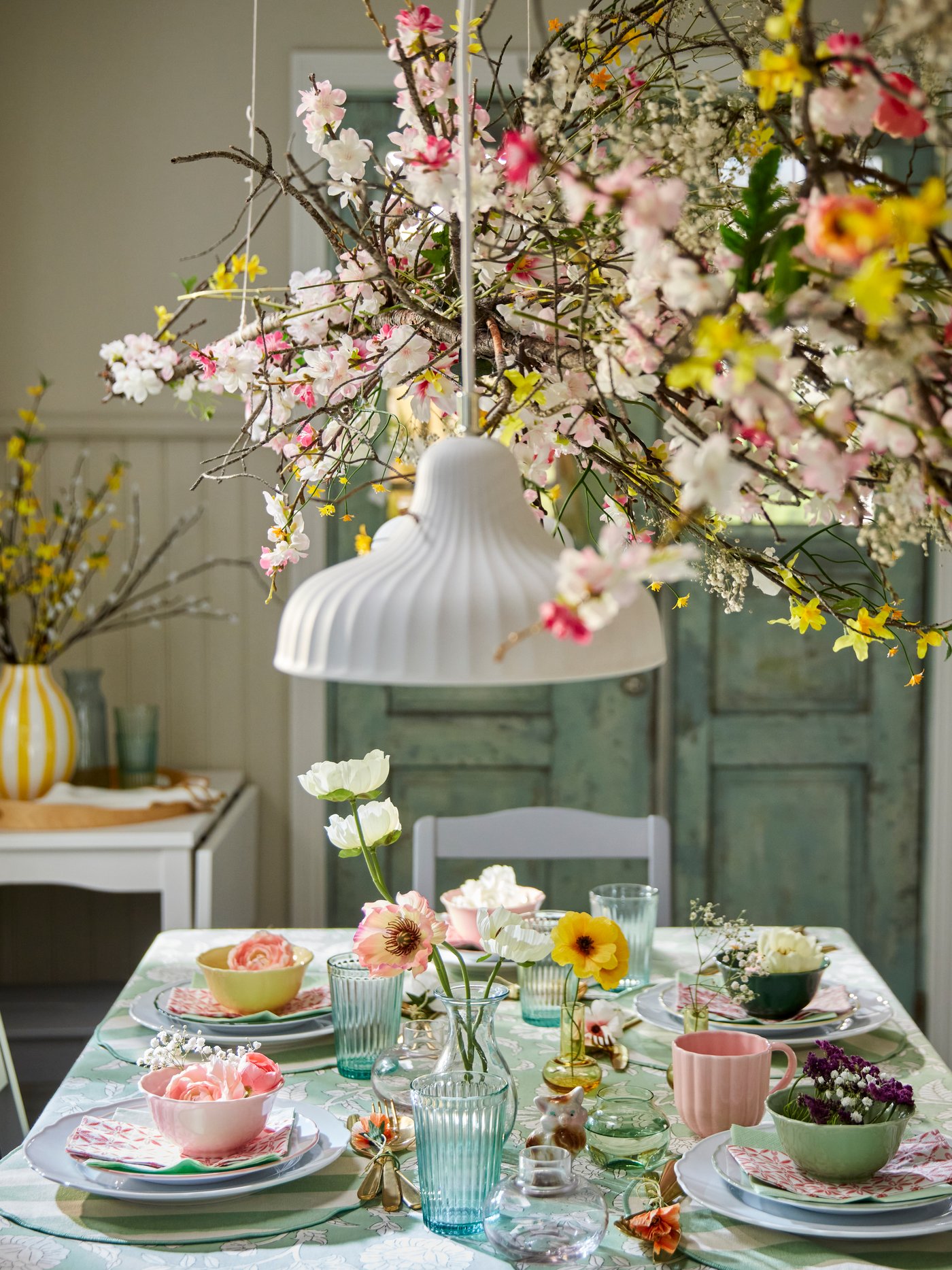 Some colourful SMÖRFISK mugs and UPPLAGA bowls sit on a beautifully set dining table adorned with flowers and greenery.