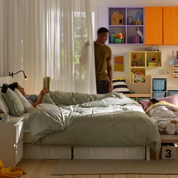 Deux personnes dans une chambre à coucher, l'une allongée dans un lit aux draps vert clair, lisant un livre, l'autre debout derrière les rideaux colorés.