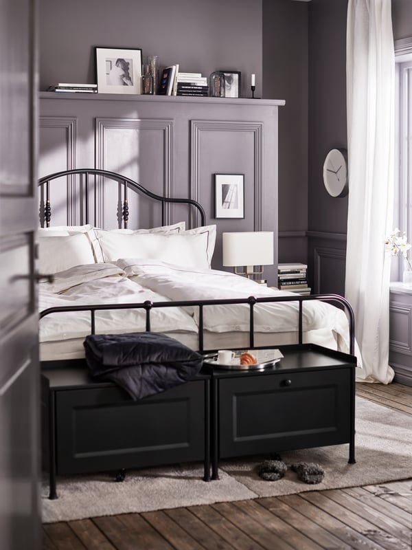 Two black KORNSJÖ storage benches and a bed with a black bed frame made with white/dark gray bed linen on a light gray rug.