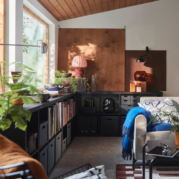 A living room features a black-brown KALLAX shelving unit holding books and decorative items beside a grey sofa.