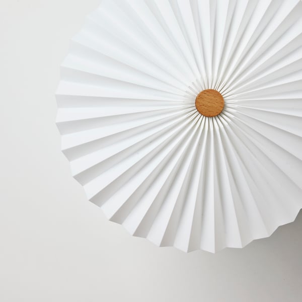 The top of a white KUDDLAVA table lamp, with light playing in the pleats of the circular shade around a central birch knob.