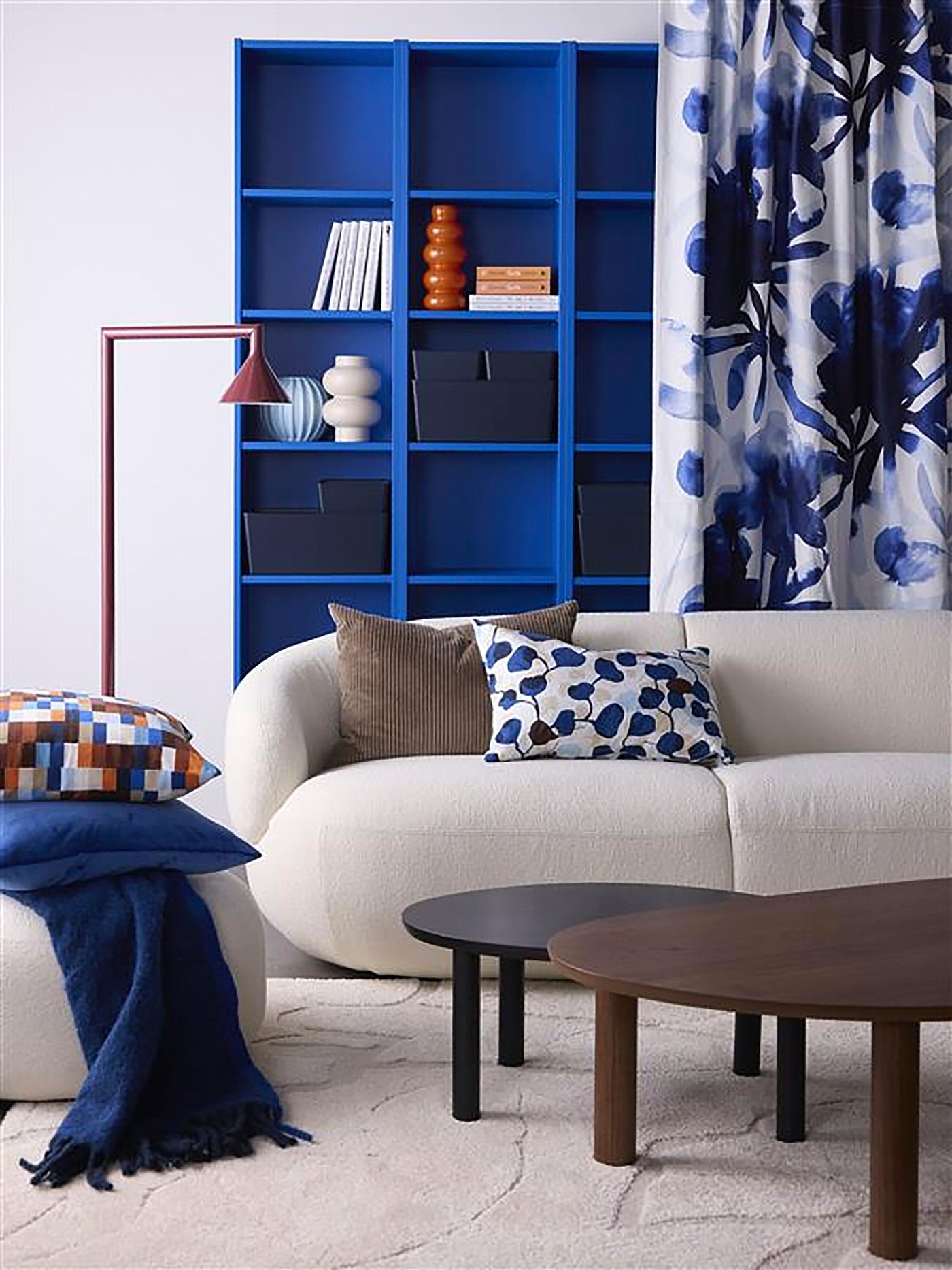 Un soggiorno con divano chiaro: sulla seduta ci sono due cuscini decorativi, uno bianco e blu e l’altro marrone. Sulla sinistra, un pouf con un cuscino con fodera LÖVNATURSKOG a quadretti colorati e un plaid blu acceso.