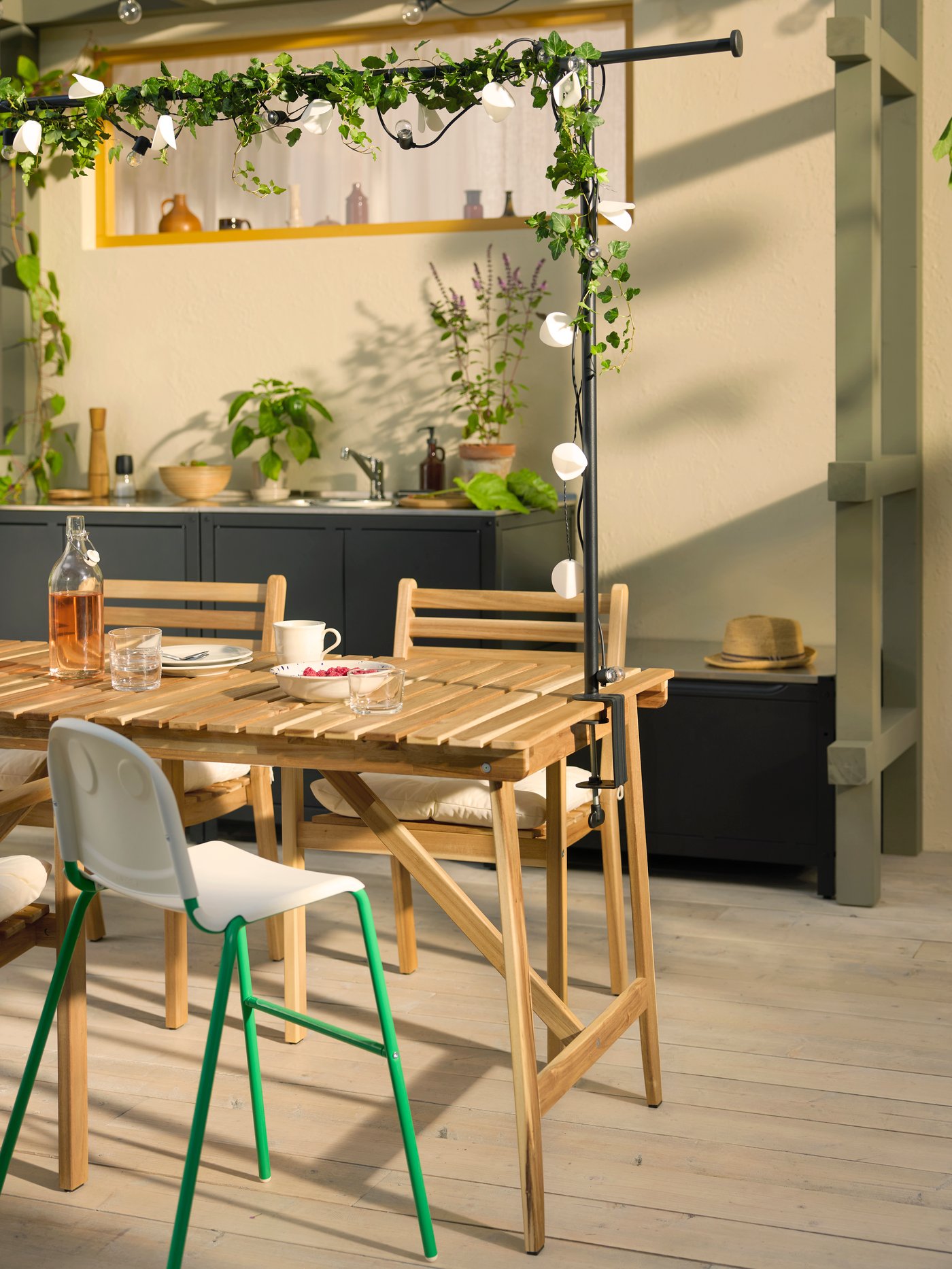 A patio with an acacia ASKHOLMEN outdoor table, outdoor chairs with armrests, and a highchair surrounded by plants.