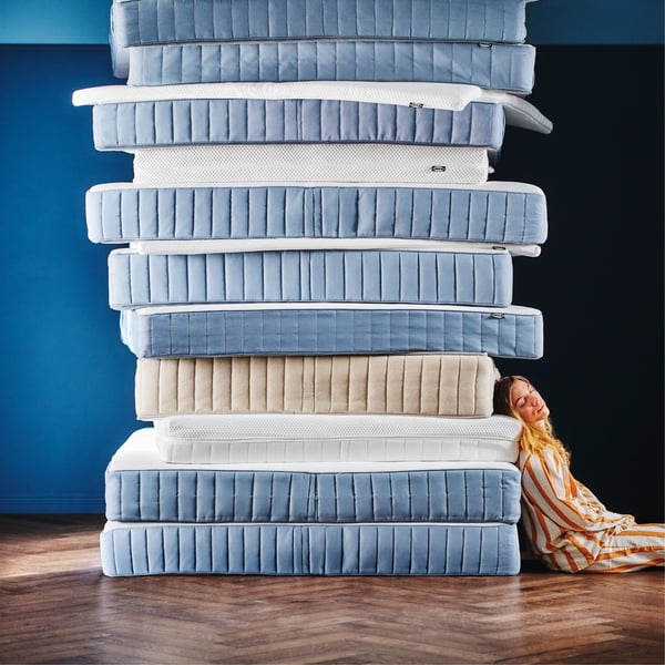 Een vrouw in gestreepte pyjama leunt tegen een hoge stapel van negen matrassen in verschillende blauwtinten en texturen, met één beige matras in het midden.