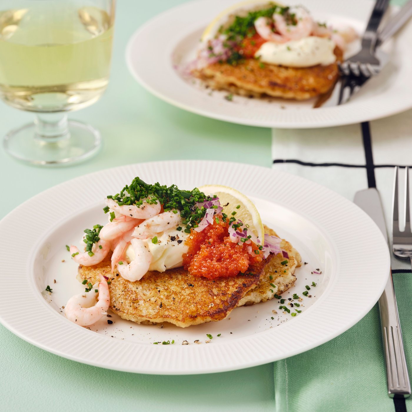 Golden-brown rösti with shrimp, caviar and a lemon slice on top are placed on two white plates on a green tablecloth.
