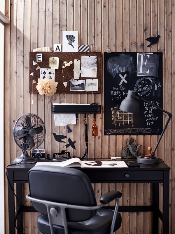 Una postazione provvista di poltrona, due bacheche di cui una di legno e una lavagna nera. Sotto le bacheche una lampada da parte, una lampada da lavoro e un ventilatore nero. Al centro della scrivania un quaderno aperto. 