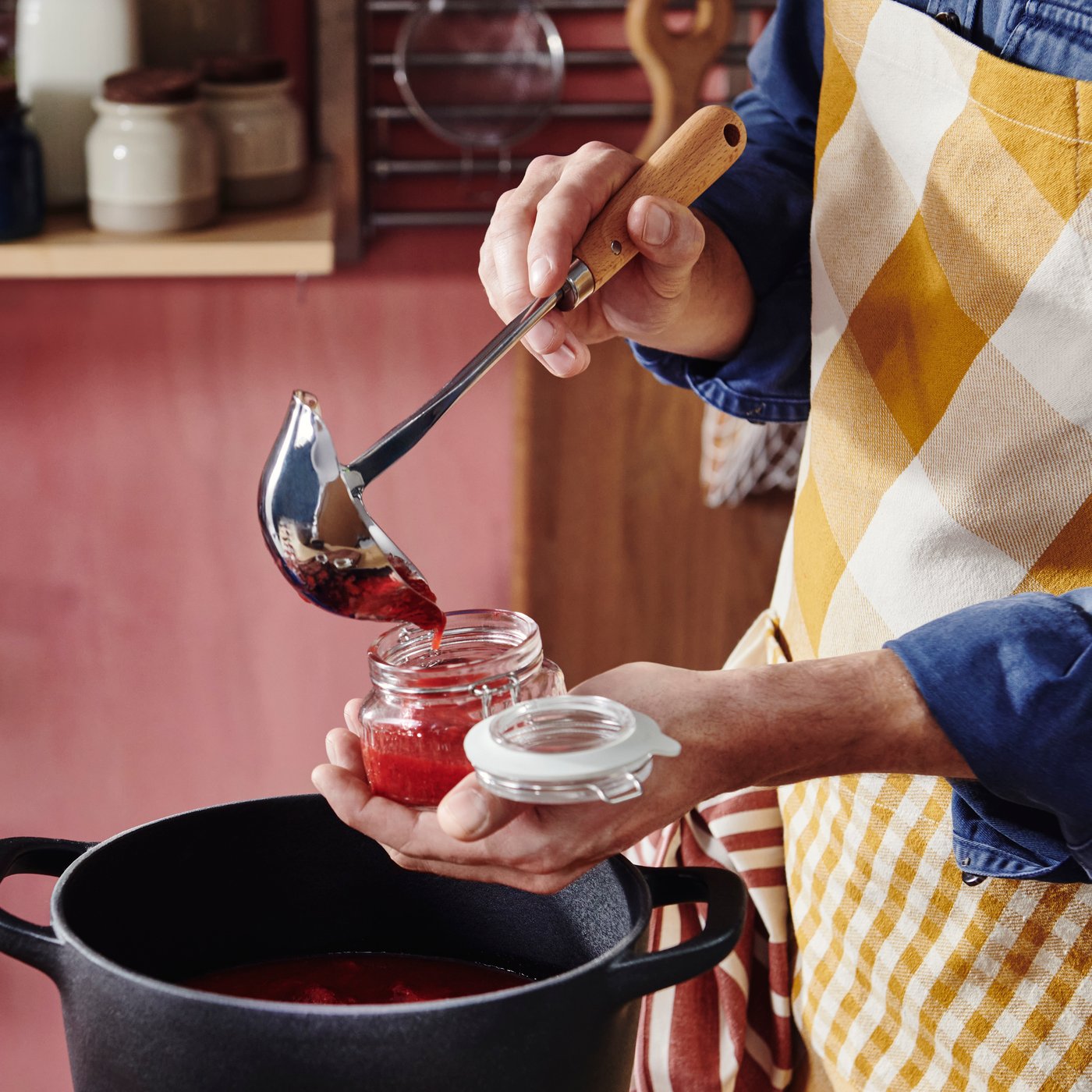A person wearing an apron holds a VARDAGEN soup ladle in one hand and a glass jar in the other, spooning jam into the jar.
