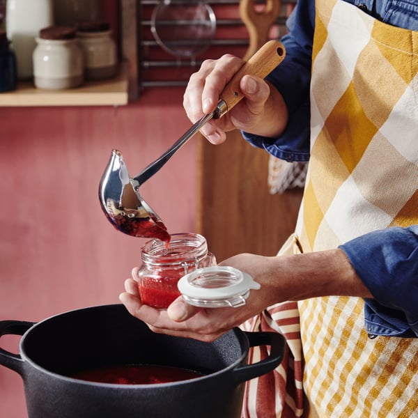 A person wearing an apron holds a VARDAGEN soup ladle in one hand and a glass jar in the other, spooning jam into the jar.