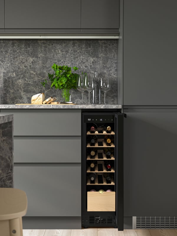 Modernt kök med grå skåp, marmorbakstycke, inbyggd SPELNÄS vinkyl med flaskor, tre vinglas, färska örter i glas och bröd på skärbräda.

