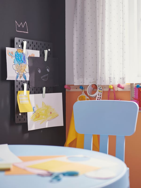 Eine schwarze SKÅDIS Lochplatte hängt in der Ecke eines Raumes an einer schwarzen Wand. An ihr sind Kinderzeichnungen mit SKÅDIS Klemmen befestigt.