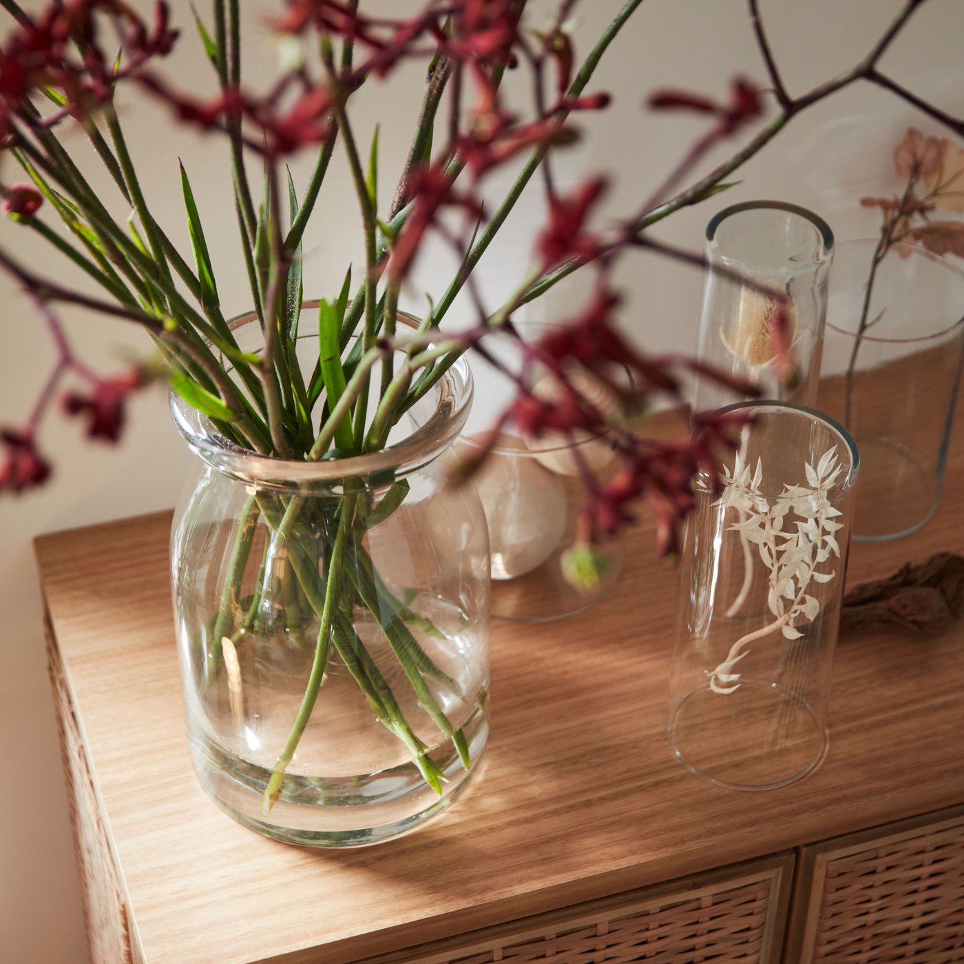 Thin flowers are loosely arranged in a clear glass BEGÄRLIG vase alongside other vases on the top of a FRYKSÅS cabinet.