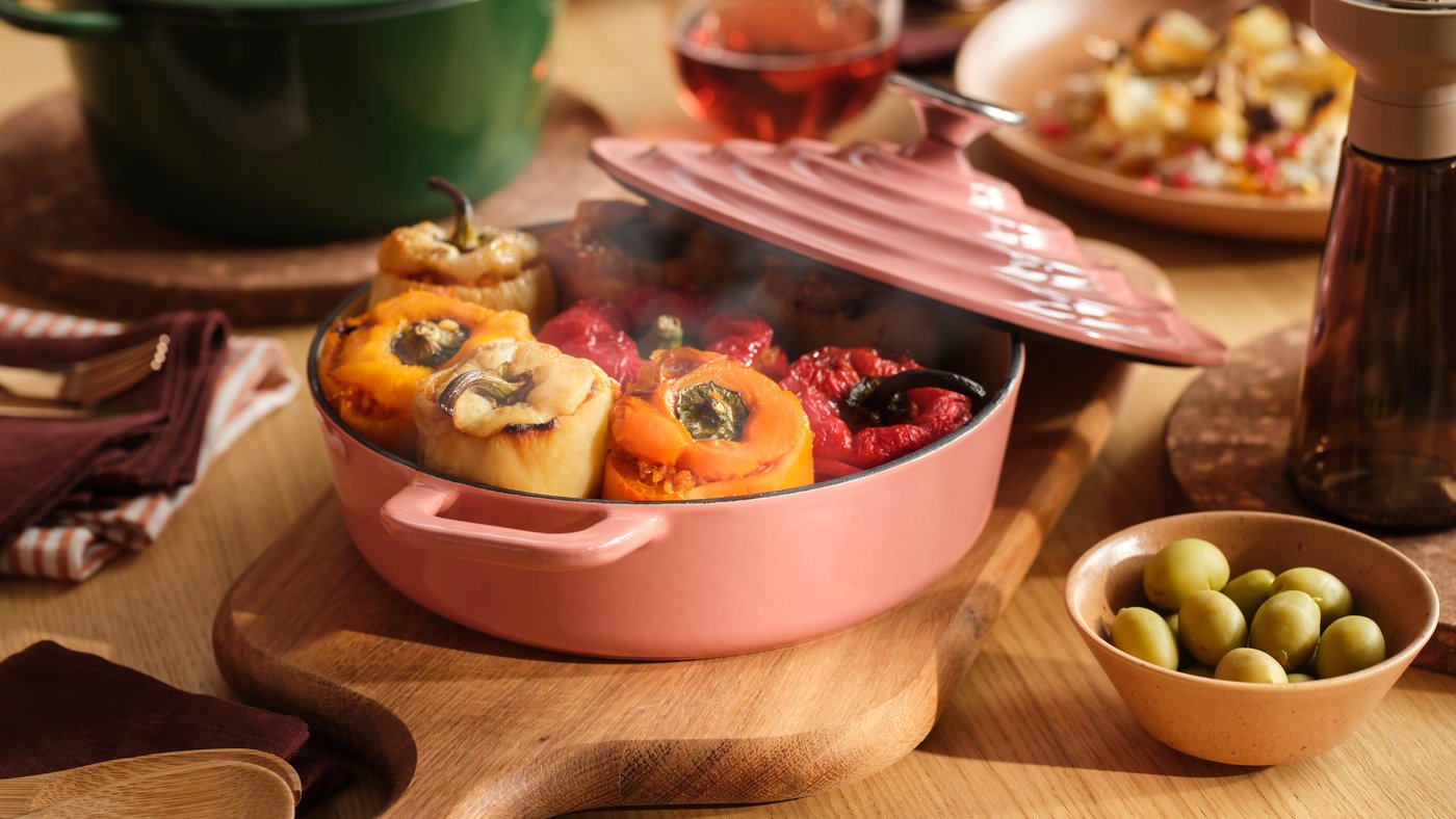 A VARDAGEN pot in light pink filled with stuffed peppers stands on a wooden dining table together with other pots and food.