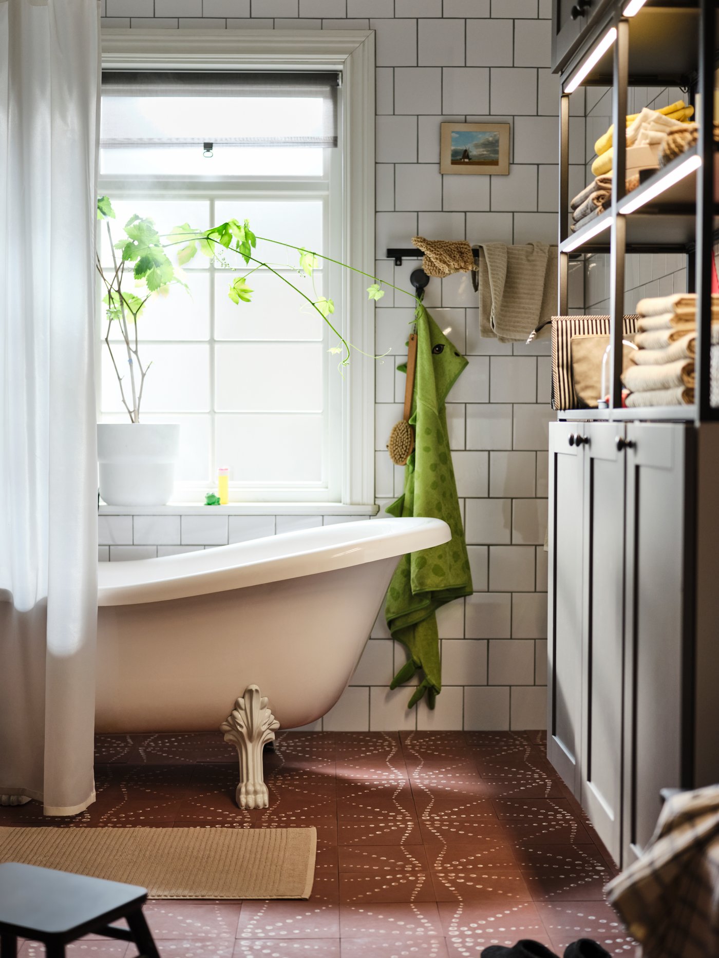 A white-tiled bathroom with a plant in the window, a bath behind a shower curtain and a grey ENHET wall storage combination.