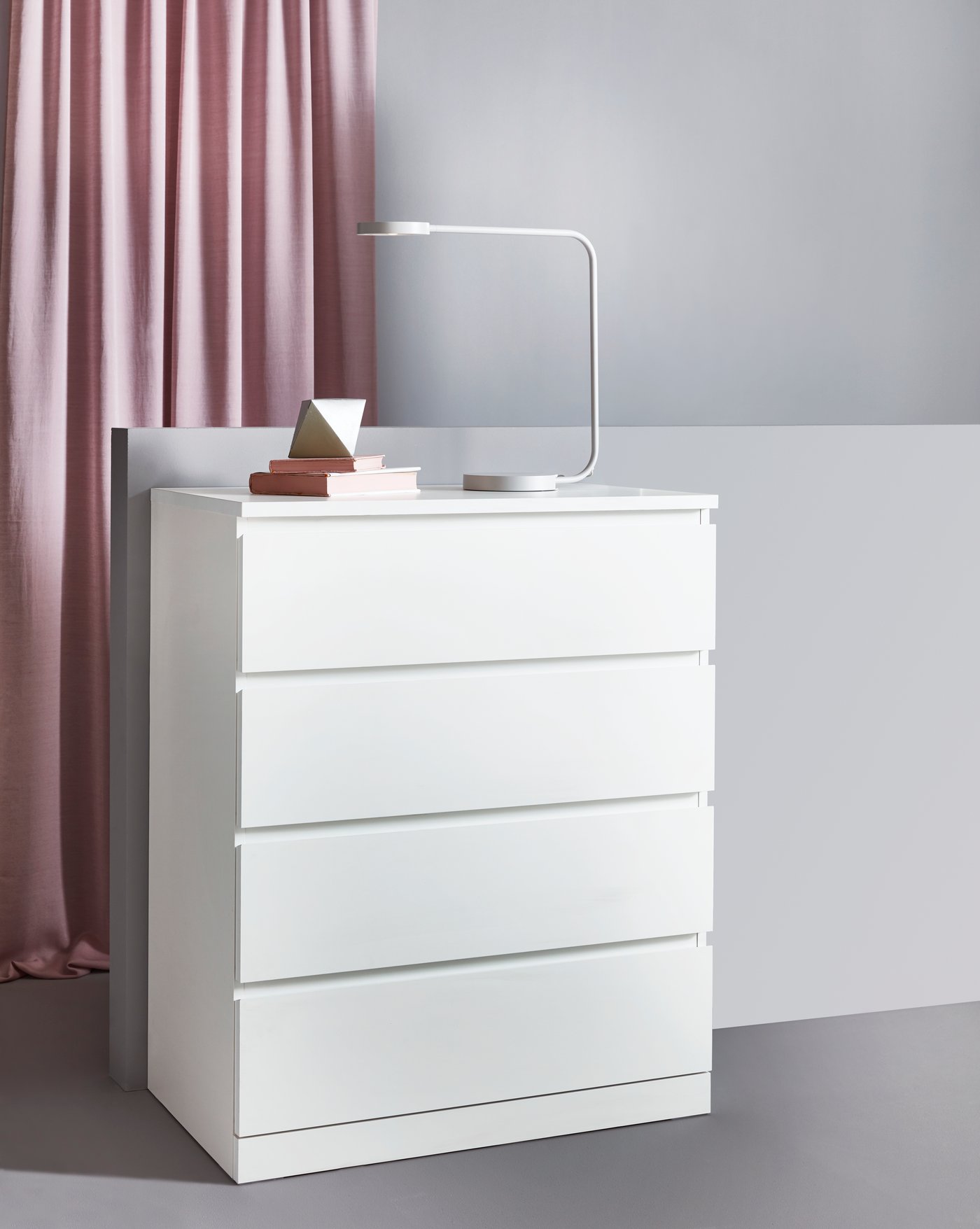 Comodă IKEA albă cu patru sertare, într-un decor minimalist, decorată cu lampă de birou și câteva cărți, pe fundal cu perdea roz – spațiu simplu și ordonat pentru depozitare de zi cu zi.