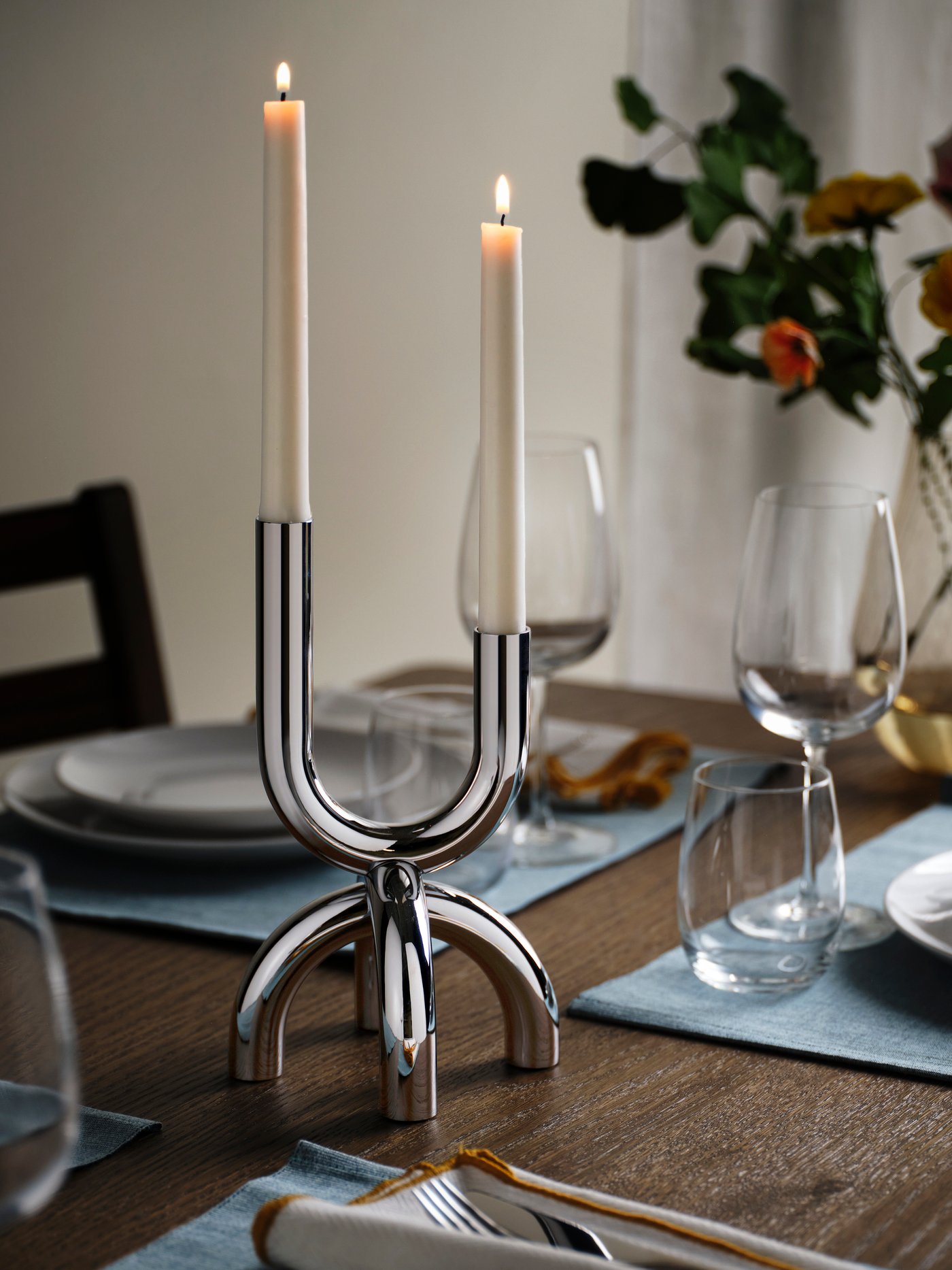 Två tända ljus i eleganta ljusstakar står på ett dukat bord med glas och en blomsterdekoration i bakgrunden.